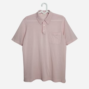 May include: A pale pink short-sleeved polo shirt with a button-up collar and a single pocket on the left chest.