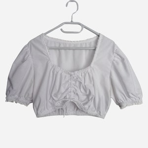 Op de afbeelding: Witte getailleerde blouse met pofmouwen en een kantversiering. De blouse heeft een geplooide halslijn en een trekkoord in de taille.