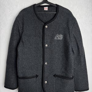 Op de afbeelding: Een grijs fleecejack met een zwarte bies en een knoopsluiting. Het jack heeft twee zakken en een logo met de tekst "M2" en daaronder de tekst "Factory Equipment".