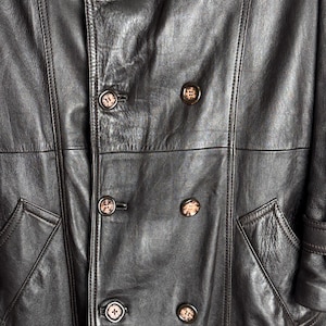 May include: Close-up of a black leather coat with a double-breasted design. The coat features large, round buttons with a mottled pattern and two front pockets. The leather has a textured appearance.