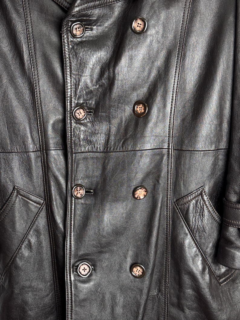 May include: Close-up of a black leather coat with a double-breasted design. The coat features large, round buttons with a mottled pattern and two front pockets. The leather has a textured appearance.