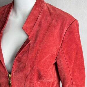 Puede incluir: Chaqueta de ante roja con cuello con solapas y cierre de cremallera dorada. La chaqueta presenta un corte entallado con costuras a lo largo de los hombros y las mangas. La chaqueta se exhibe en un maniqu&iacute;.