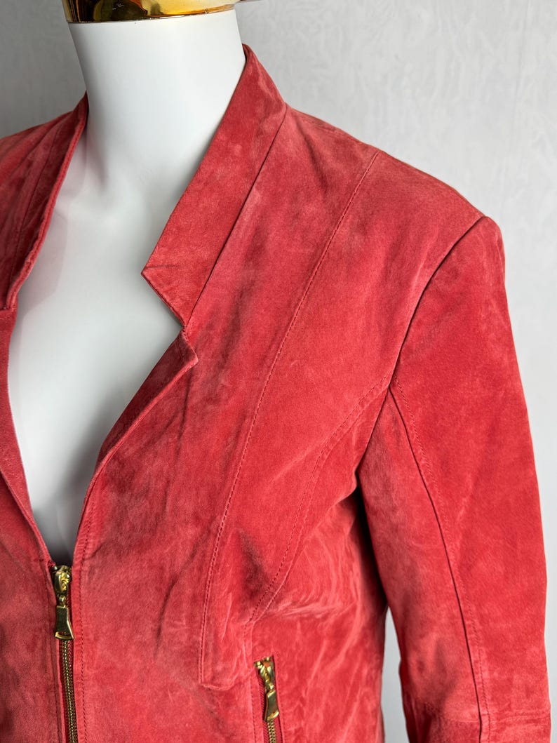 Puede incluir: Chaqueta de ante roja con cuello con solapas y cierre de cremallera dorada. La chaqueta presenta un corte entallado con costuras a lo largo de los hombros y las mangas. La chaqueta se exhibe en un maniqu&iacute;.