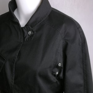 May include: A black cotton jacket with a collared neckline, a button closure, and a belt. The jacket has a slightly fitted silhouette and features a patch pocket on the left side.