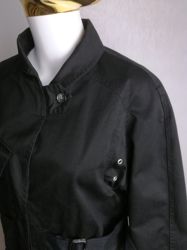 May include: A black cotton jacket with a collared neckline, a button closure, and a belt. The jacket has a slightly fitted silhouette and features a patch pocket on the left side.