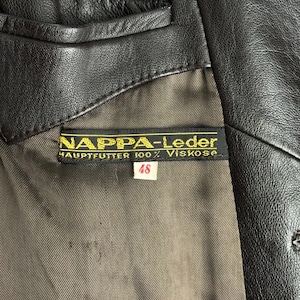 May include: Close-up of a black leather jacket with a label that reads "NAPPA-Leder". The label also indicates "HAUPTFUTTER 100% Viskose" and size "48". The jacket has a brown lining.