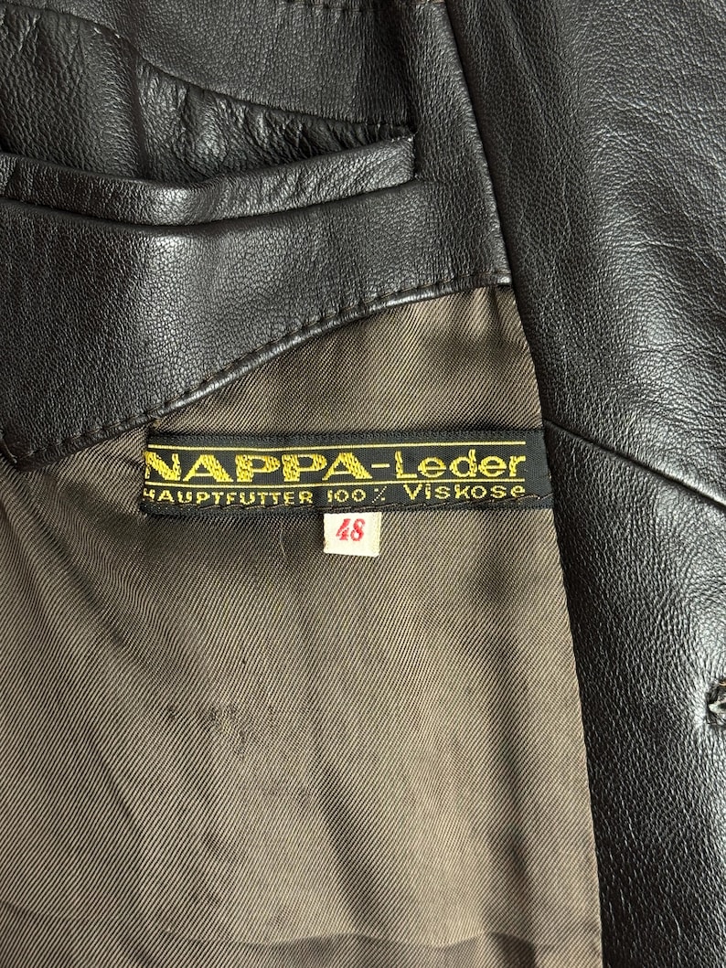 May include: Close-up of a black leather jacket with a label that reads "NAPPA-Leder". The label also indicates "HAUPTFUTTER 100% Viskose" and size "48". The jacket has a brown lining.