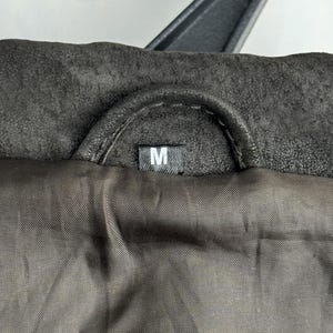 Puede incluir: Primer plano de un cuello de chaqueta marr&oacute;n oscuro con una percha negra. El forro interior es de un marr&oacute;n m&aacute;s oscuro. Una peque&ntilde;a etiqueta blanca con la letra "M" es visible.