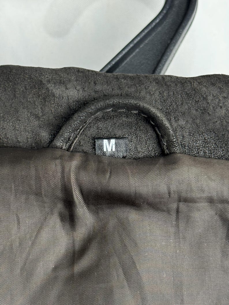 Puede incluir: Primer plano de un cuello de chaqueta marr&oacute;n oscuro con una percha negra. El forro interior es de un marr&oacute;n m&aacute;s oscuro. Una peque&ntilde;a etiqueta blanca con la letra "M" es visible.