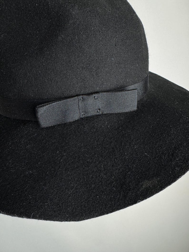 Puede incluir: Un sombrero de fieltro negro de ala ancha con una cinta de grosgrain negra y un lazo.