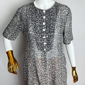 May include: A sheer, short-sleeved blouse with a dark gray background and a white floral pattern. The blouse has a round neckline and a row of white buttons down the front. The bottom of the blouse is paired with a light pink pleated skirt.
