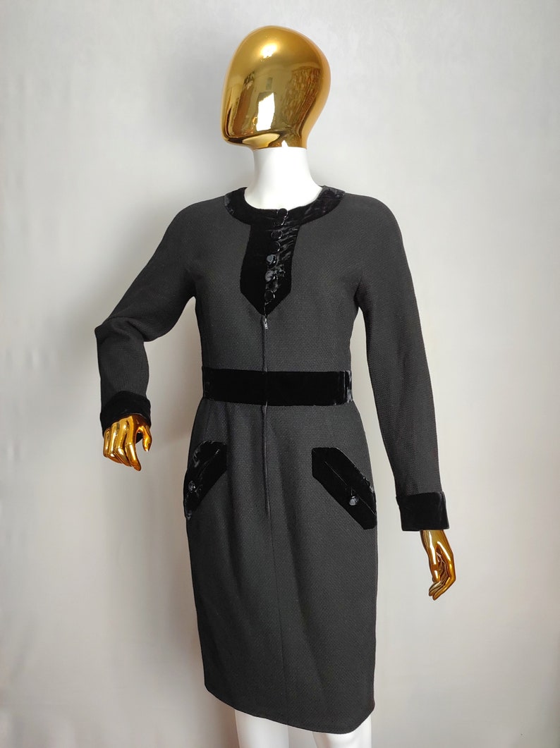 K&ouml;nnte beinhalten: Ein schwarzes Kleid mit Samtbesatz an Ausschnitt, Taille und &Auml;rmelb&uuml;ndchen. Das Kleid hat einen Rei&szlig;verschluss und zwei Taschen mit Samtbesatz.