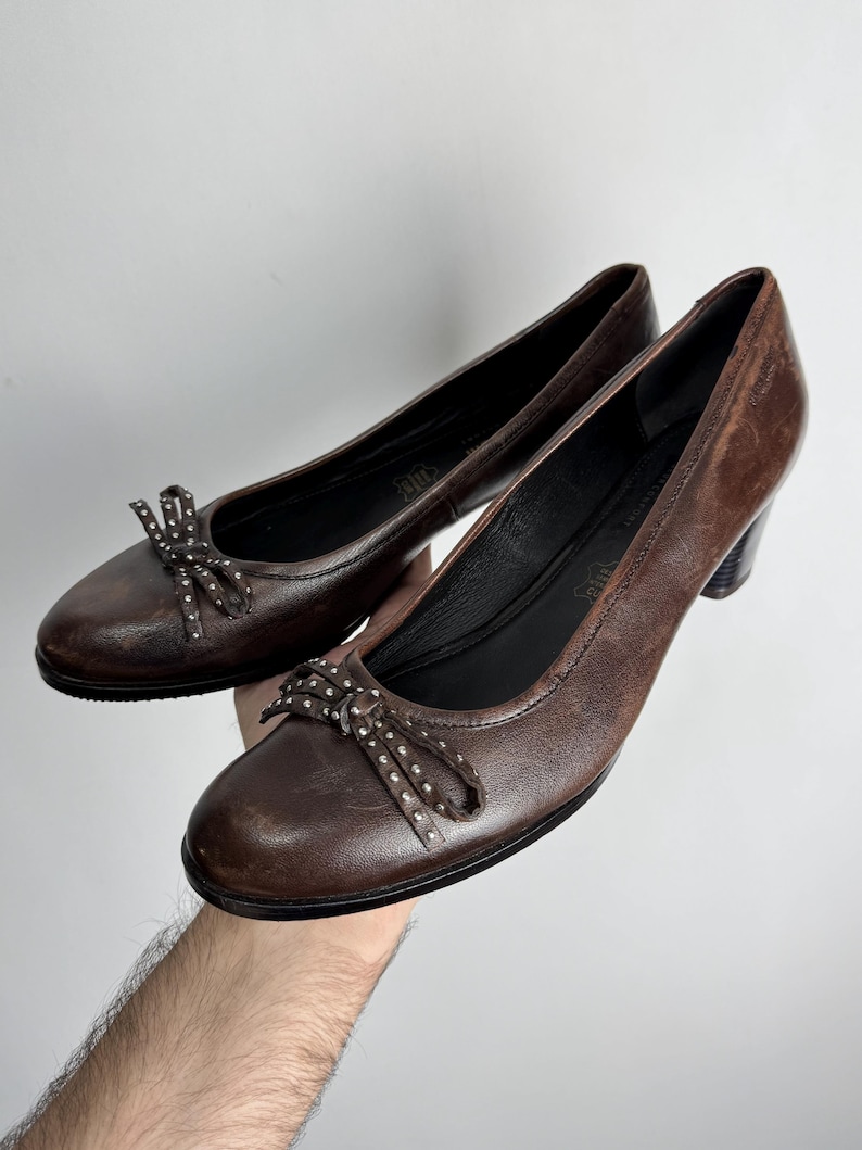 Puede incluir: Un par de zapatos de sal&oacute;n de cuero marr&oacute;n con peque&ntilde;os lazos adornados con tachuelas plateadas. Los zapatos tienen una punta redondeada y un tac&oacute;n peque&ntilde;o. El cuero tiene un aspecto ligeramente envejecido.