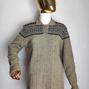 May include: A long-sleeved, collared sweater in a neutral beige color. The sweater features a patterned design across the shoulders in shades of blue, green, and black. The ribbed knit fabric and button-up collar add texture and detail to the garment.