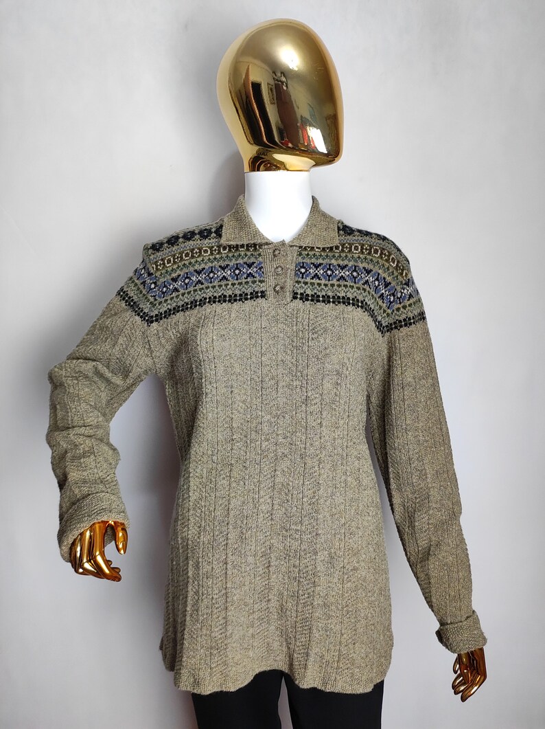 May include: A long-sleeved, collared sweater in a neutral beige color. The sweater features a patterned design across the shoulders in shades of blue, green, and black. The ribbed knit fabric and button-up collar add texture and detail to the garment.