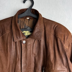 May include: A brown leather jacket with a collared neckline and a button closure. The jacket has a pocket on the chest and the text "CAVAL CON" is visible on the label. The jacket is hanging on a black hanger.
