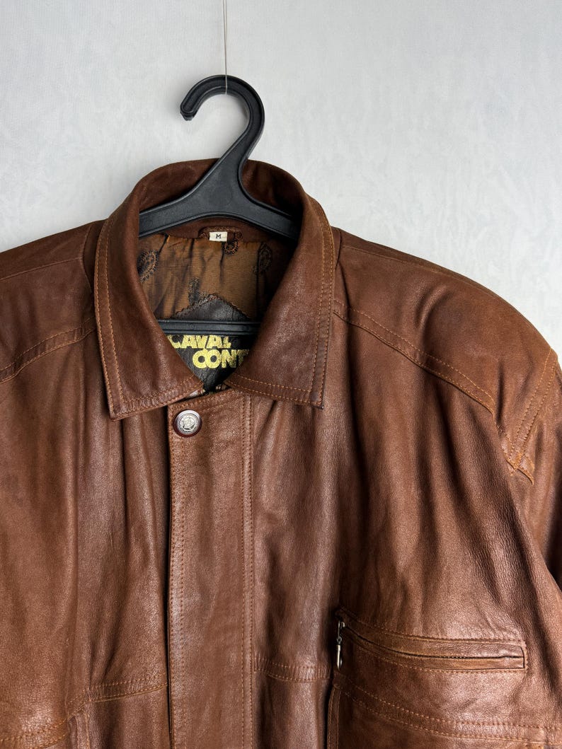 May include: A brown leather jacket with a collared neckline and a button closure. The jacket has a pocket on the chest and the text "CAVAL CON" is visible on the label. The jacket is hanging on a black hanger.
