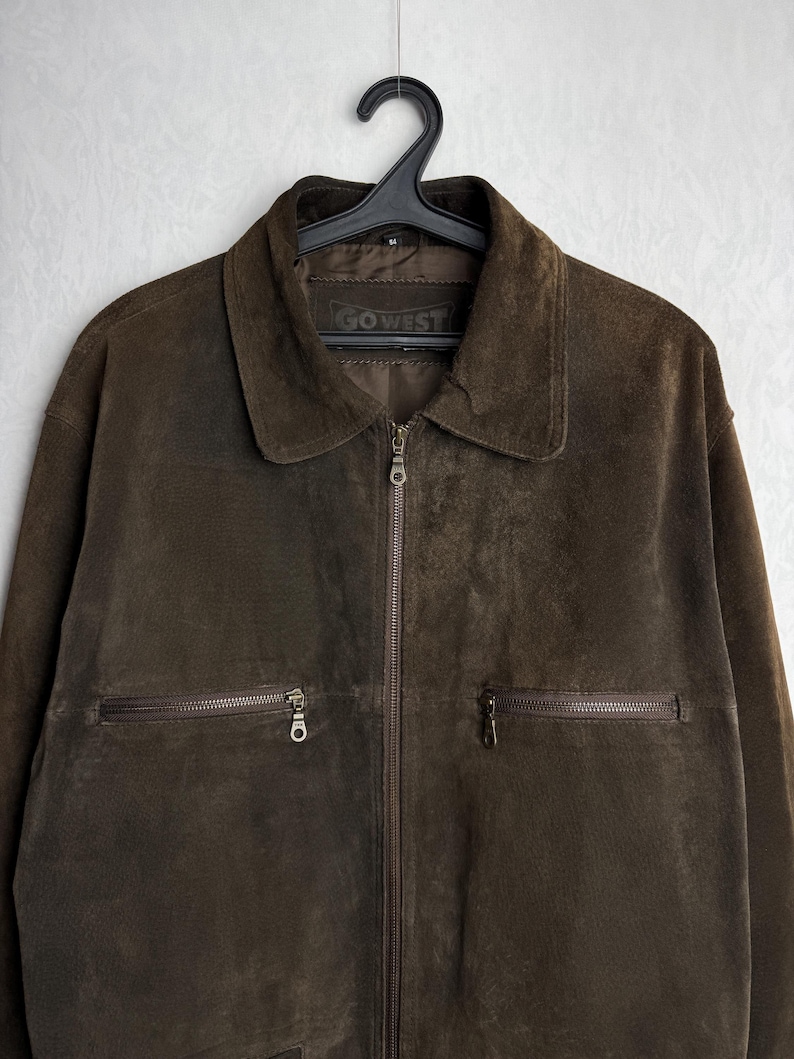 May include: A brown suede jacket with a zipper closure and two zippered pockets. The jacket has a label that reads "GO WEST".