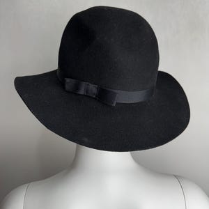 Puede incluir: Un sombrero fedora de fieltro negro con ala ancha y una cinta de sat&eacute;n negro atada en un lazo en la parte delantera.