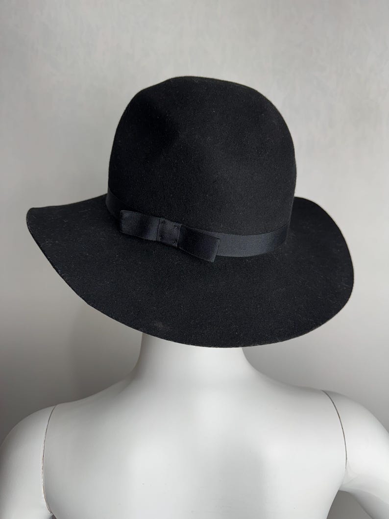 Puede incluir: Un sombrero fedora de fieltro negro con ala ancha y una cinta de sat&eacute;n negro atada en un lazo en la parte delantera.