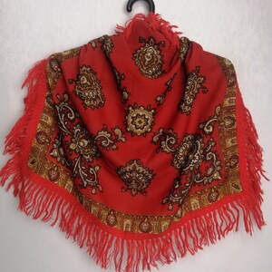 K&ouml;nnte beinhalten: Ein roter Schal mit Paisley-Muster und Fransenbesatz. Der Schal h&auml;ngt an einem schwarzen Kleiderb&uuml;gel vor einem wei&szlig;en Hintergrund.