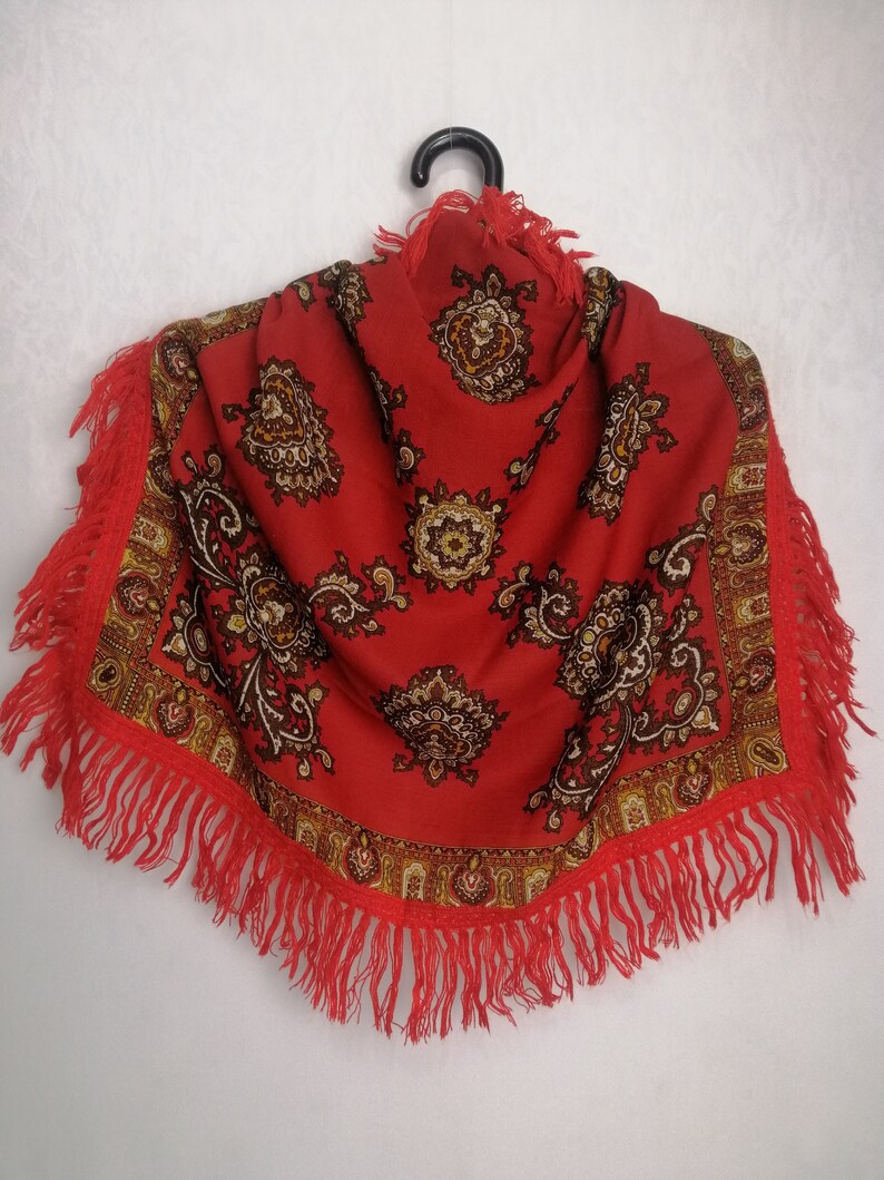 K&ouml;nnte beinhalten: Ein roter Schal mit Paisley-Muster und Fransenbesatz. Der Schal h&auml;ngt an einem schwarzen Kleiderb&uuml;gel vor einem wei&szlig;en Hintergrund.
