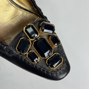 Puede incluir: Primer plano de un zapato de cuero negro con interior dorado. El zapato est&aacute; adornado con piedras preciosas rectangulares negras engastadas en marcos dorados. La costura alrededor de las piedras es visible.