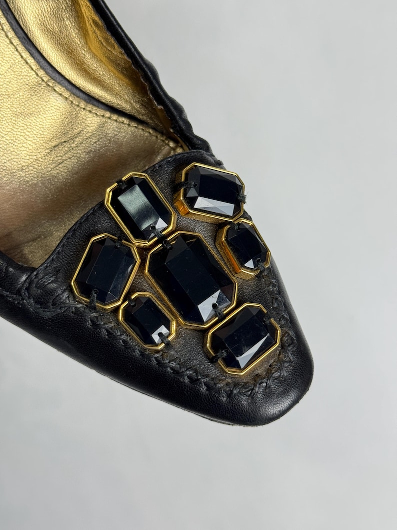 Puede incluir: Primer plano de un zapato de cuero negro con interior dorado. El zapato est&aacute; adornado con piedras preciosas rectangulares negras engastadas en marcos dorados. La costura alrededor de las piedras es visible.