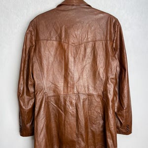 May include: A brown leather jacket hanging on a black hanger. The jacket has a classic design with a notched collar, buttoned cuffs, and a tailored fit. The leather appears to be well-worn, with a rich, textured appearance.