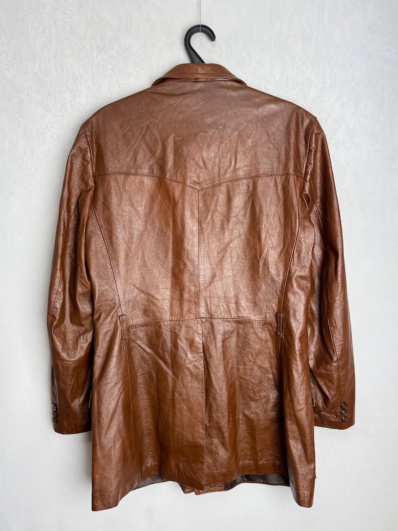 May include: A brown leather jacket hanging on a black hanger. The jacket has a classic design with a notched collar, buttoned cuffs, and a tailored fit. The leather appears to be well-worn, with a rich, textured appearance.