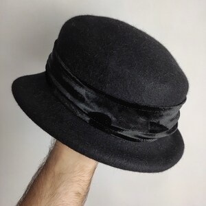 May include: A black cloche hat with a velvet band. The hat has a rounded crown and a small, downward-sloping brim. The velvet band encircles the crown, adding a touch of elegance to the classic design. This hat is a stylish accessory.