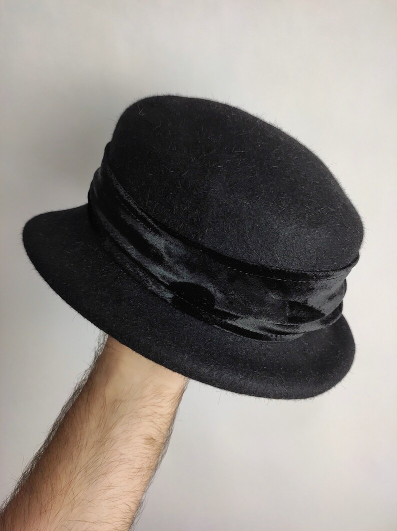 May include: A black cloche hat with a velvet band. The hat has a rounded crown and a small, downward-sloping brim. The velvet band encircles the crown, adding a touch of elegance to the classic design. This hat is a stylish accessory.