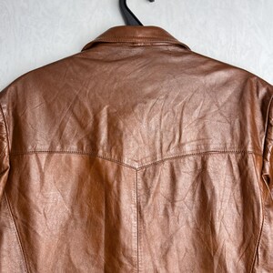 May include: A brown leather jacket hangs from a black hanger against a white background. The jacket has a classic collar and a textured surface. The back of the jacket is visible, showcasing the stitching details and design.