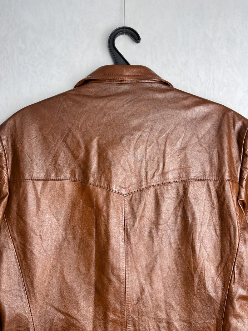 May include: A brown leather jacket hangs from a black hanger against a white background. The jacket has a classic collar and a textured surface. The back of the jacket is visible, showcasing the stitching details and design.