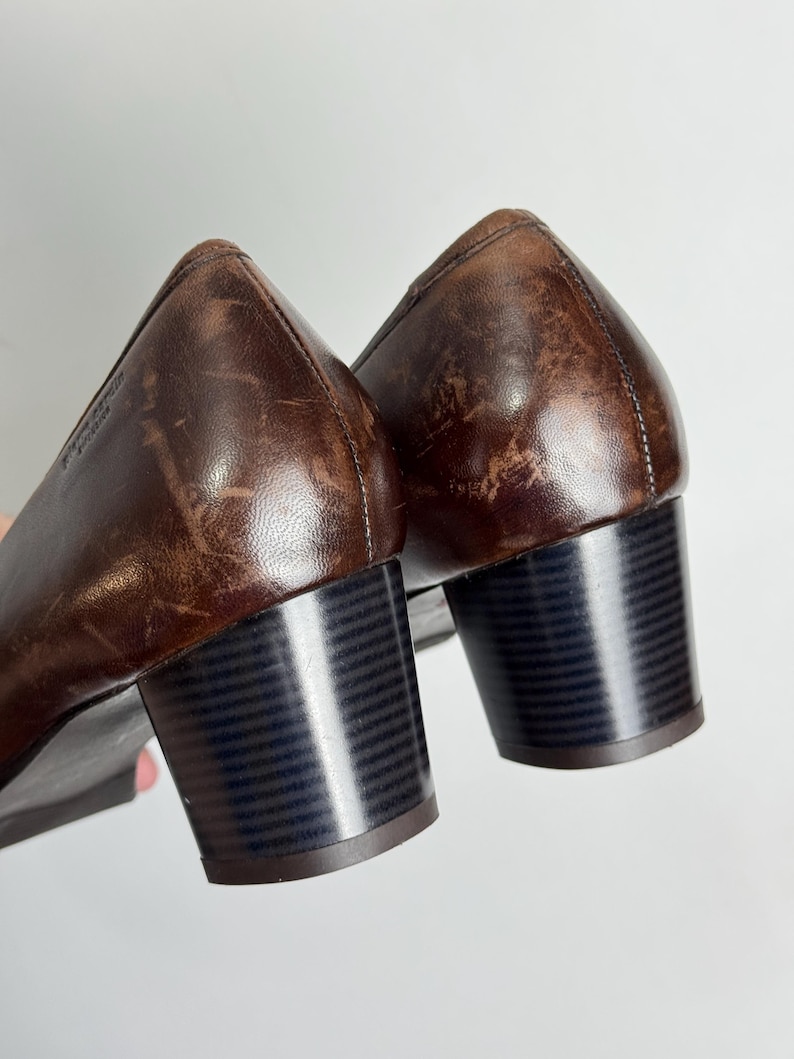 Puede incluir: Un par de zapatos de tac&oacute;n de cuero marr&oacute;n. Los zapatos tienen un tac&oacute;n de bloque y una punta redondeada. El cuero tiene un aspecto desgastado. Los tacones miden aproximadamente 5 cm de alto.