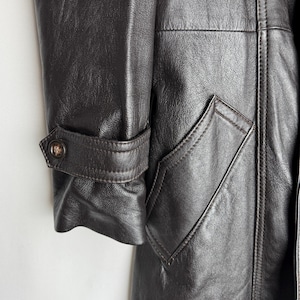 May include: Close-up of a dark brown leather coat. The coat features a buttoned cuff detail and a pocket with a pointed flap. The leather has a textured appearance, and the stitching is visible.
