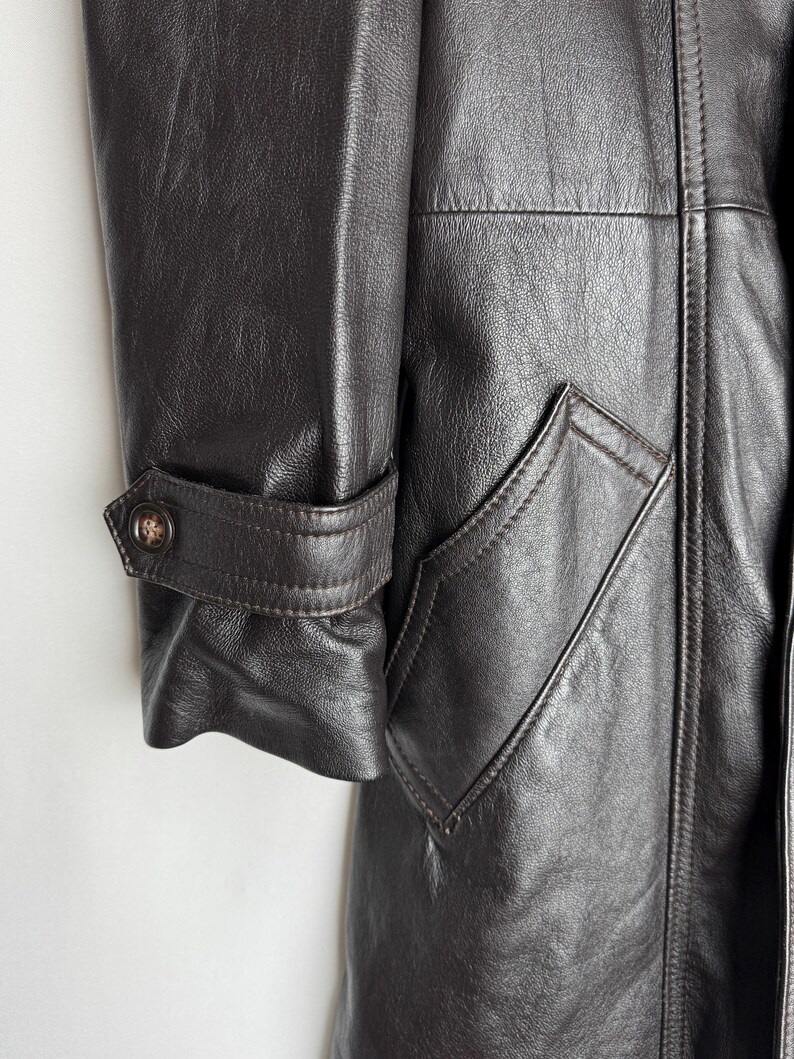 May include: Close-up of a dark brown leather coat. The coat features a buttoned cuff detail and a pocket with a pointed flap. The leather has a textured appearance, and the stitching is visible.