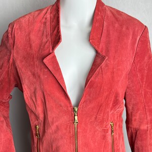 Puede incluir: Chaqueta de ante color coral con cuello de solapa y cremallera dorada completa. La chaqueta tiene dos bolsillos con cremallera y un corte entallado, expuesta en un maniqu&iacute;.
