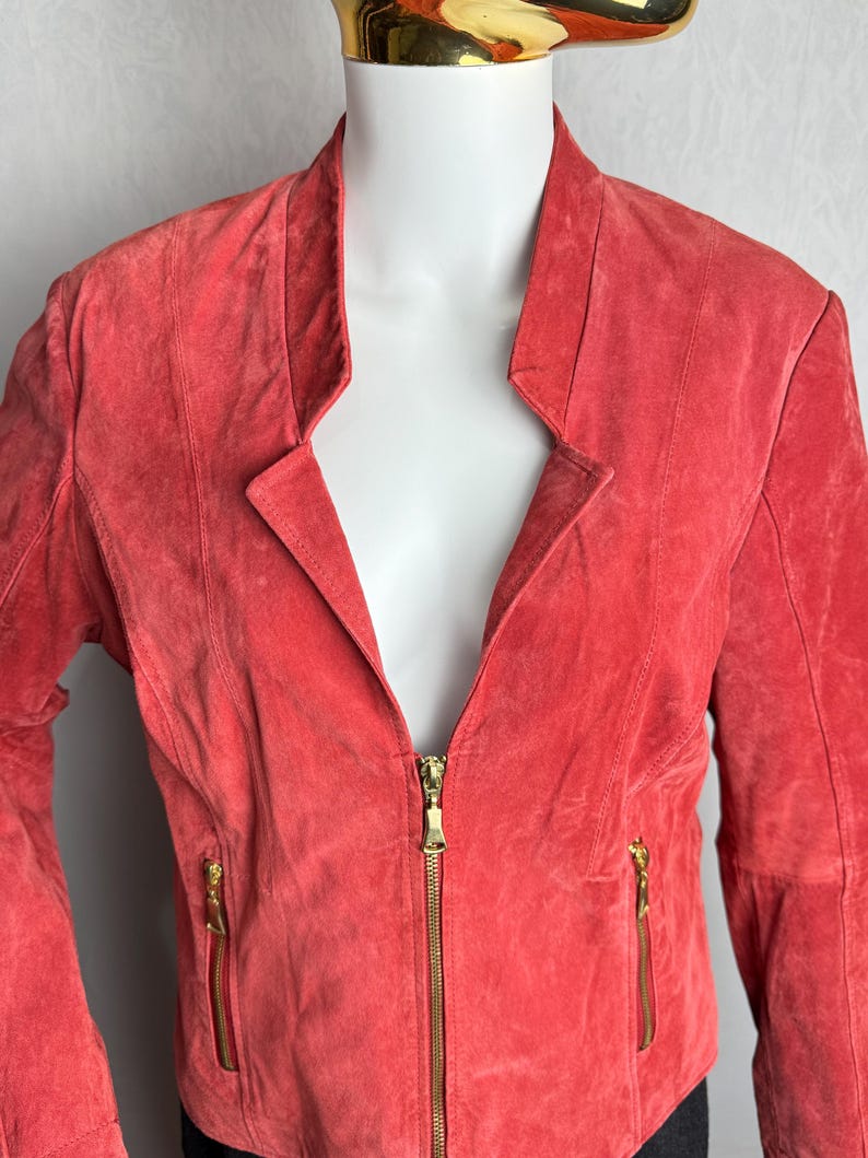Puede incluir: Chaqueta de ante color coral con cuello de solapa y cremallera dorada completa. La chaqueta tiene dos bolsillos con cremallera y un corte entallado, expuesta en un maniqu&iacute;.