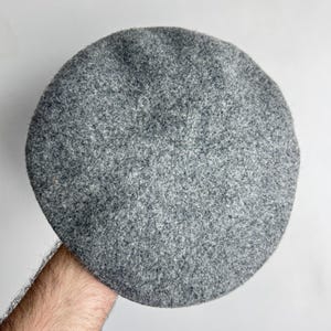 Pode incluir: Um boina cinzenta feita de um tecido texturizado. O chap&eacute;u &eacute; redondo e tem um design cl&aacute;ssico. A boina &eacute; de uma cor neutra, adequada para v&aacute;rias roupas e estilos.