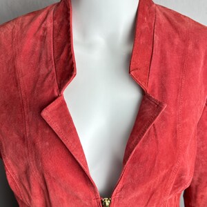 Puede incluir: Chaqueta de ante color coral con cuello con solapas y cremallera dorada. La chaqueta tiene un corte entallado con costuras verticales y cremalleras doradas en las mangas. La chaqueta se exhibe en un maniqu&iacute; blanco.
