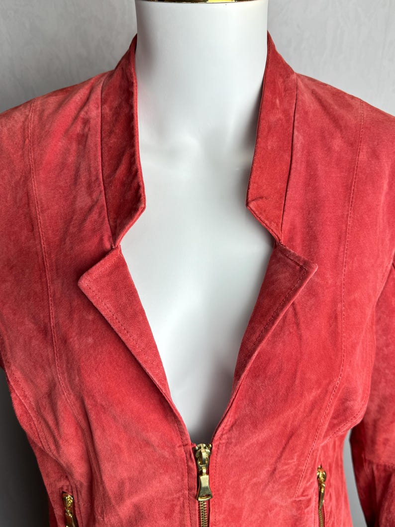 Puede incluir: Chaqueta de ante color coral con cuello con solapas y cremallera dorada. La chaqueta tiene un corte entallado con costuras verticales y cremalleras doradas en las mangas. La chaqueta se exhibe en un maniqu&iacute; blanco.