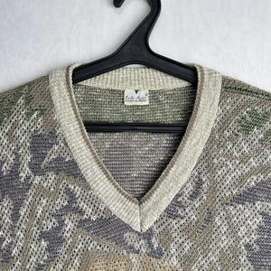 May include: A vintage V-neck sweater in shades of green, gray, and beige, hanging on a black hanger. The sweater features a patterned design and a ribbed neckline. The label reads "Emilio Valenti".