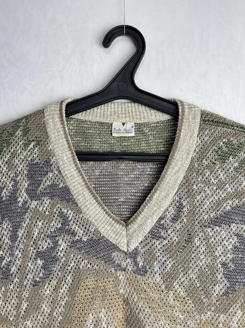May include: A vintage V-neck sweater in shades of green, gray, and beige, hanging on a black hanger. The sweater features a patterned design and a ribbed neckline. The label reads "Emilio Valenti".