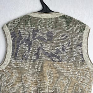 May include: A sleeveless sweater with a patterned design in shades of green, gray, beige, and burgundy. The sweater is made of a knit material and has a ribbed neckline and armholes. The garment is hanging on a black hanger.