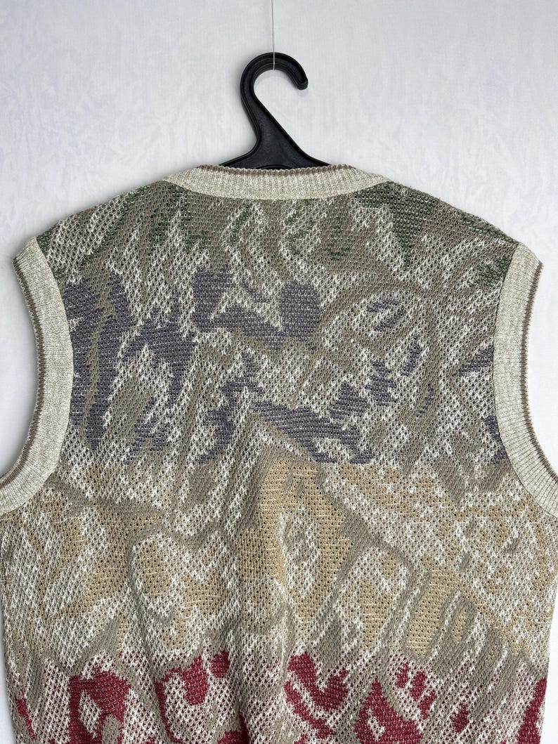 May include: A sleeveless sweater with a patterned design in shades of green, gray, beige, and burgundy. The sweater is made of a knit material and has a ribbed neckline and armholes. The garment is hanging on a black hanger.