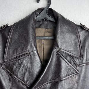 May include: A dark brown leather jacket with a double-breasted design, hanging on a black hanger. The jacket features a wide collar, button closures, and shoulder epaulettes. The leather appears worn and textured.