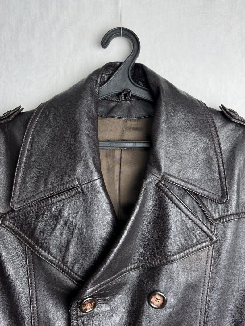 May include: A dark brown leather jacket with a double-breasted design, hanging on a black hanger. The jacket features a wide collar, button closures, and shoulder epaulettes. The leather appears worn and textured.