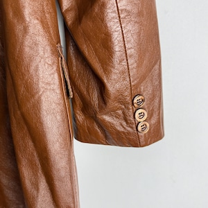 May include: A close-up of a brown leather coat. The coat has a classic design with a notched lapel and three wooden buttons on the sleeve. The leather has a textured appearance.