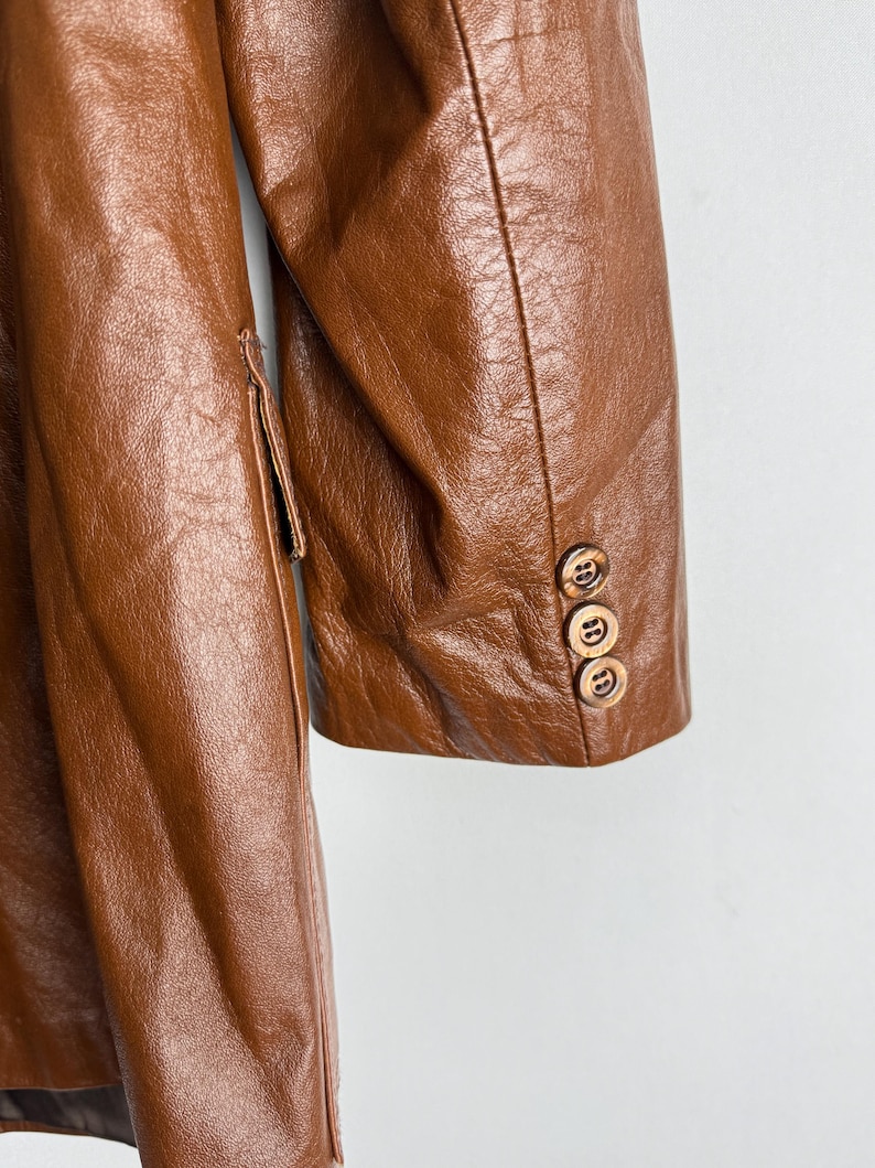 May include: A close-up of a brown leather coat. The coat has a classic design with a notched lapel and three wooden buttons on the sleeve. The leather has a textured appearance.